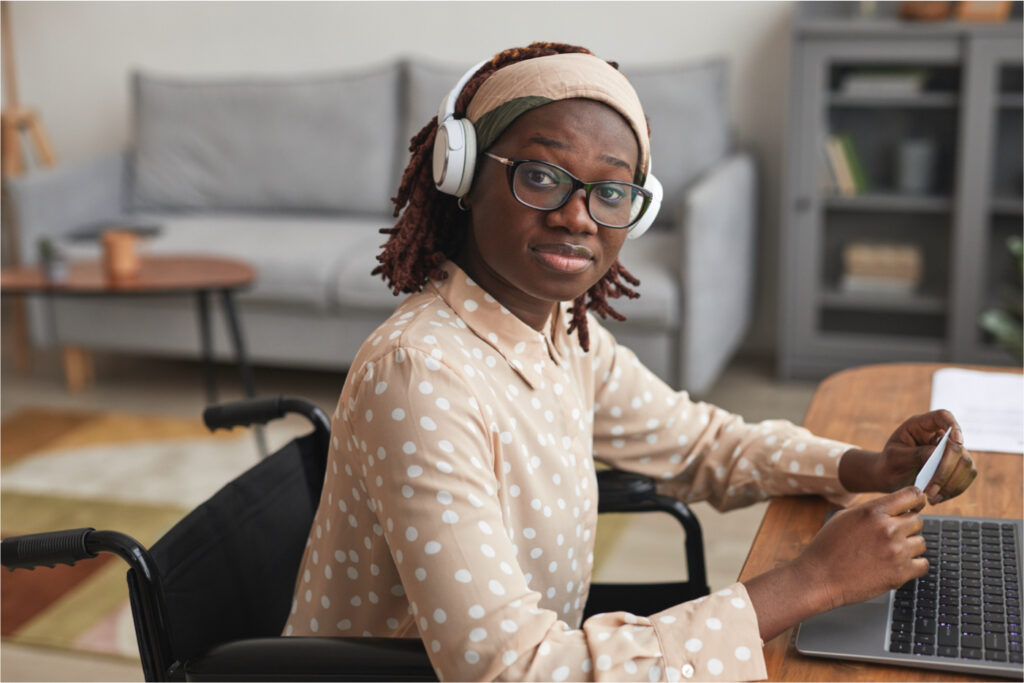 A person with dark brown skin smiling in a tan and white polka dot shirt wearing glasses and white headphones sitting in a black wheelchair typing with a lounge space blurred in the background.