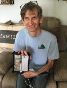 Image of a person with fair skin with gray and brown hair grinning while holding an assistive technology device