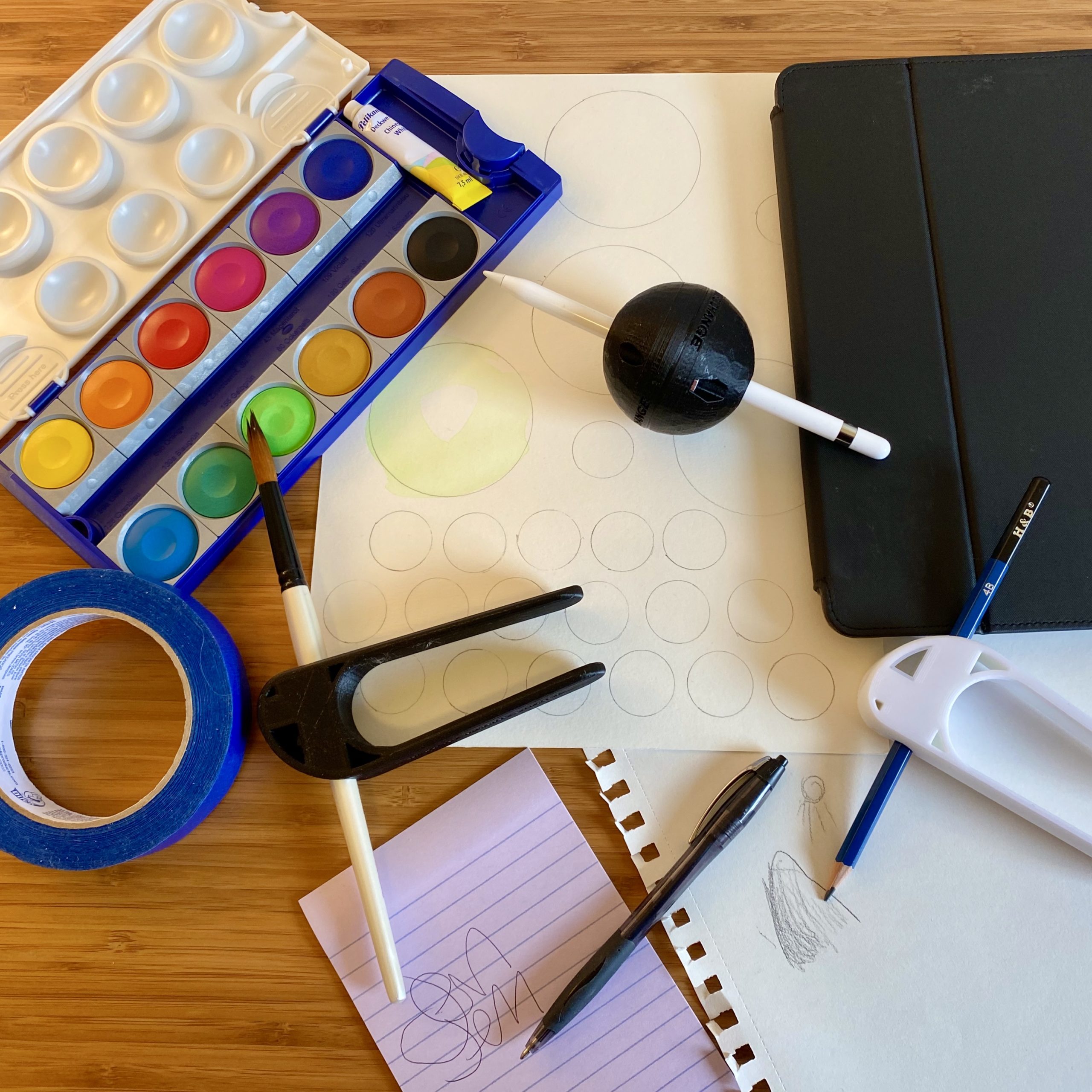AT-for-Writing-Painting-and-Drawing-scaled A watercolor paint palette, a Pen Ball device holding an Apple Pencil, a piece of watercolor paper, an iPad, a Palm Pen Holder device holding a blue drawing pencil pointing to a piece of sketch paper and small sketch of a hill and sunshine, a lined piece of paper with writing on it next to a pen, another Palm Pen Holder holding a paintbrush, and a roll of blue painters tape. All of the items are on top of a wooden desk and the photo was taken from above.