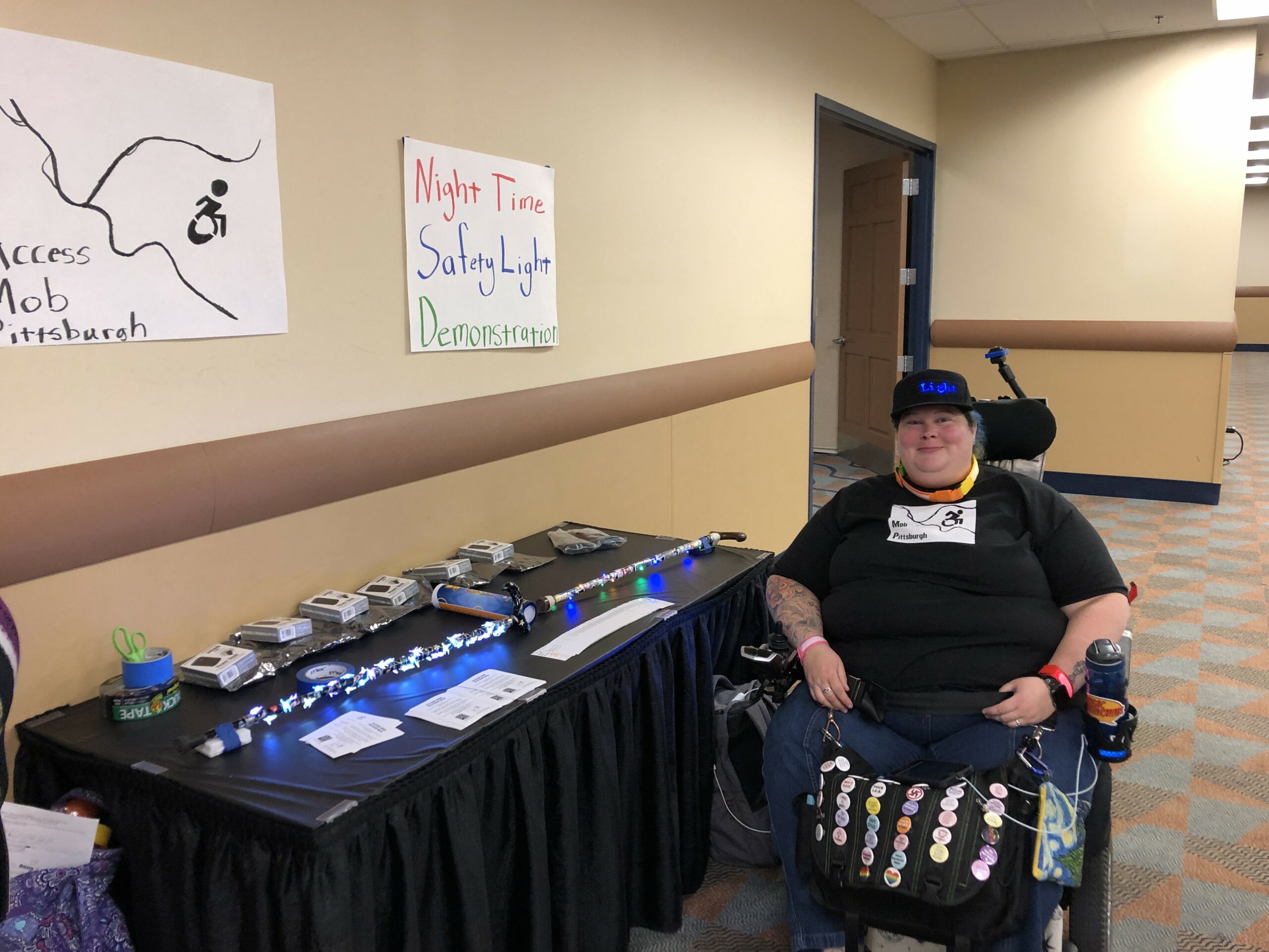 Alisa Alisa Grishman seated in her power chair next to a table that features illuminated canes. A sign for Access Mob Pittsburgh and a sign for Night Time Safety Light Demonstration hang on the wall above the table.