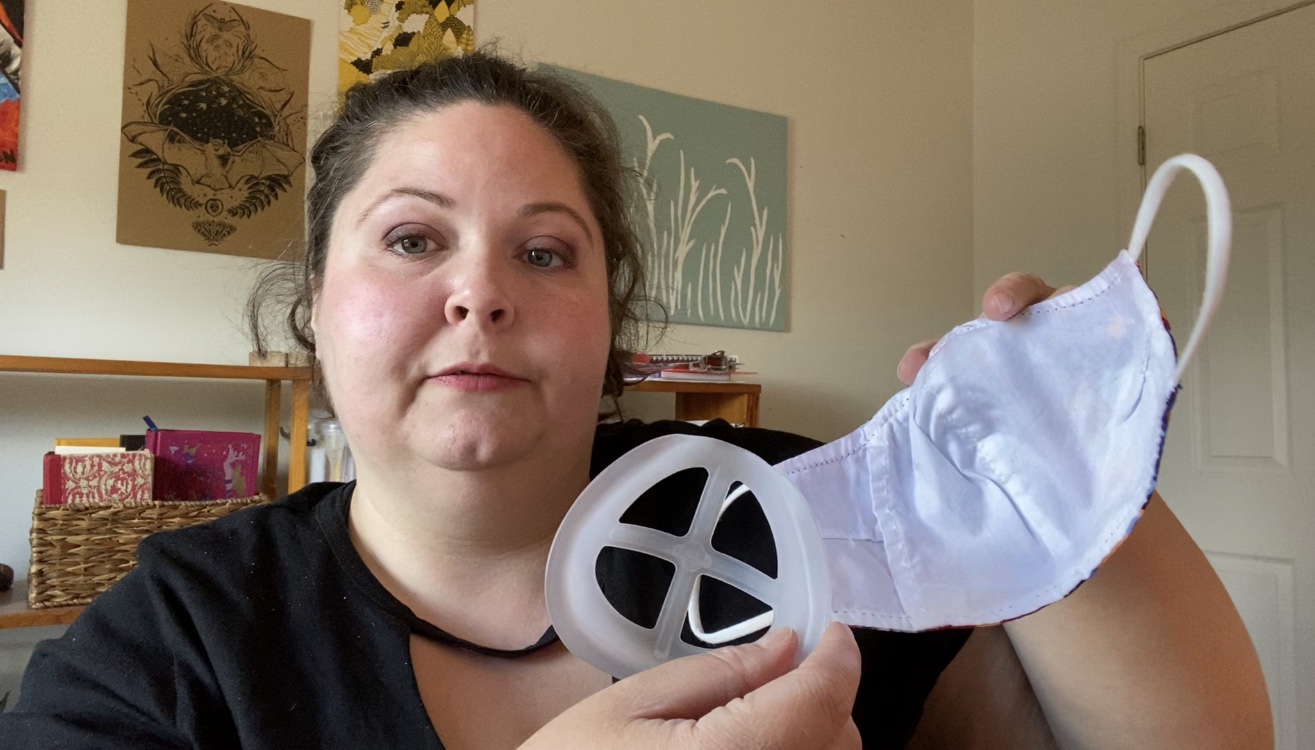 Jen Mullins is a white woman who has her wavy, brown hair up in a ponytail. Jen is holding up a face mask silicone mask insert. In the background is a wooden bookshelf, a wall with various art pieces, and a closet door.   