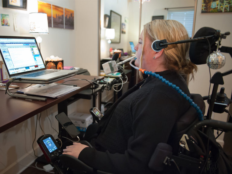Woman using AT to work hands-free with puff device control, including voice control and additional asisstive devices besides computer