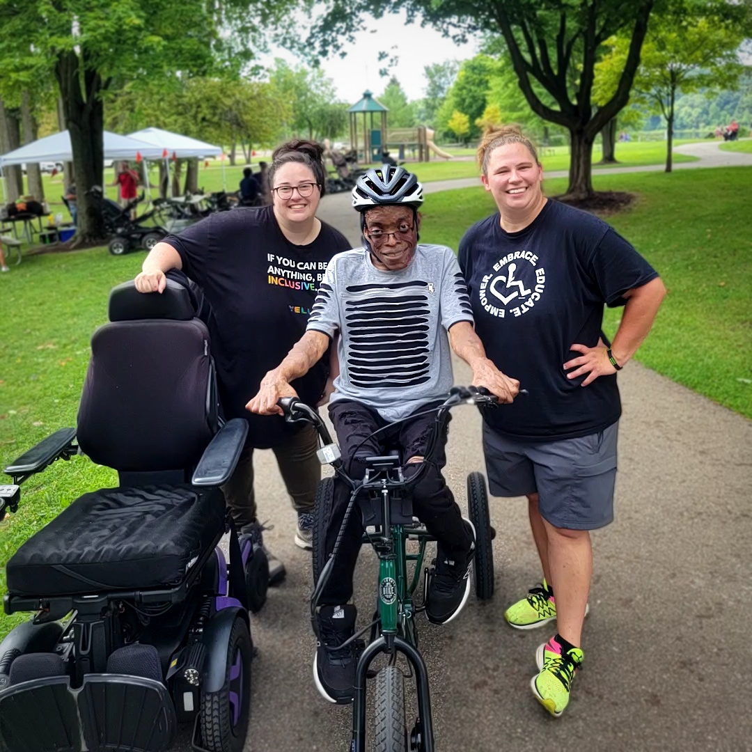 Two staff members posing with an individual wearing a helmet using adaptive bicycle in park setting