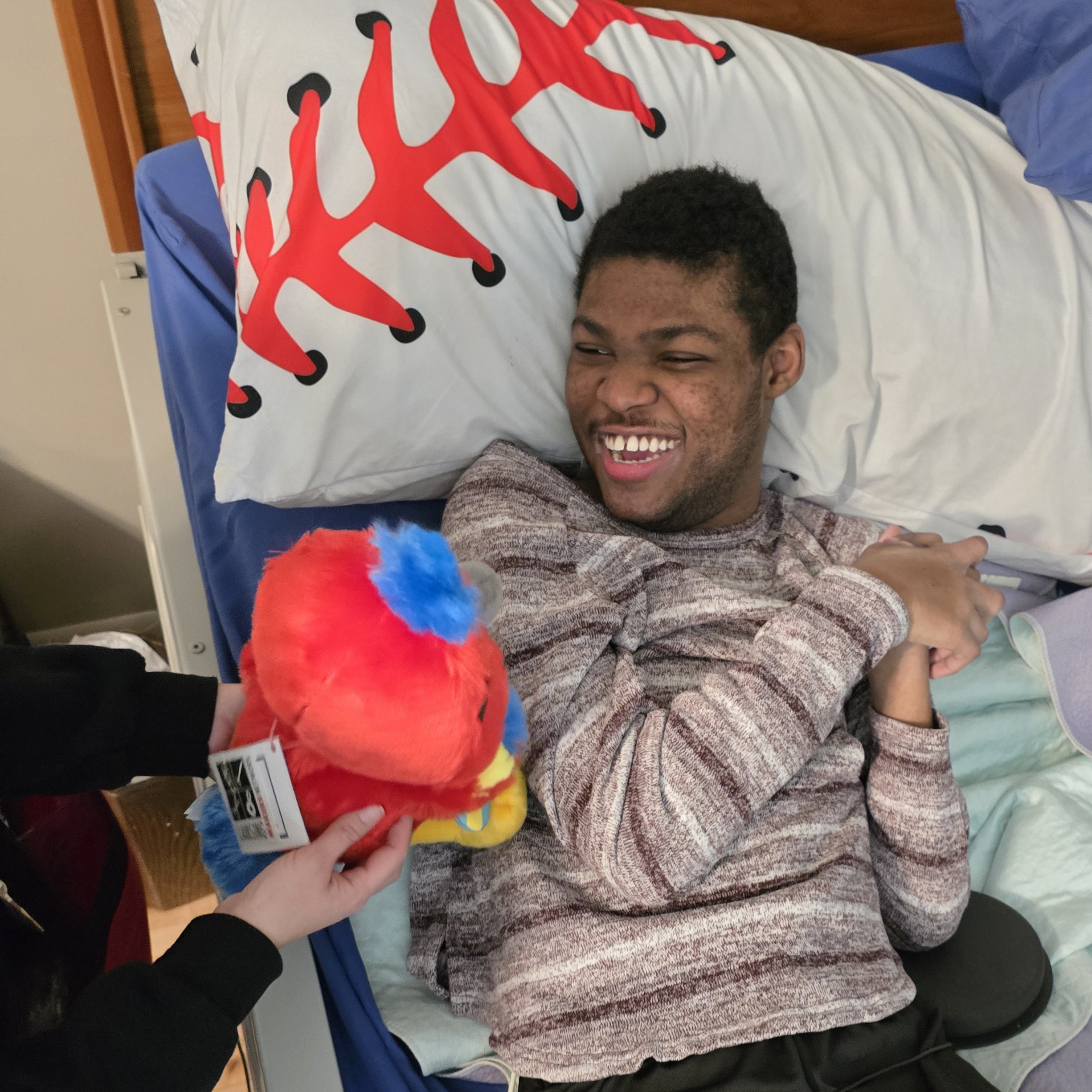 Young man smiling in bed while staff member demonstrates adaptive toy which man is operating with an elbow hitting a switch