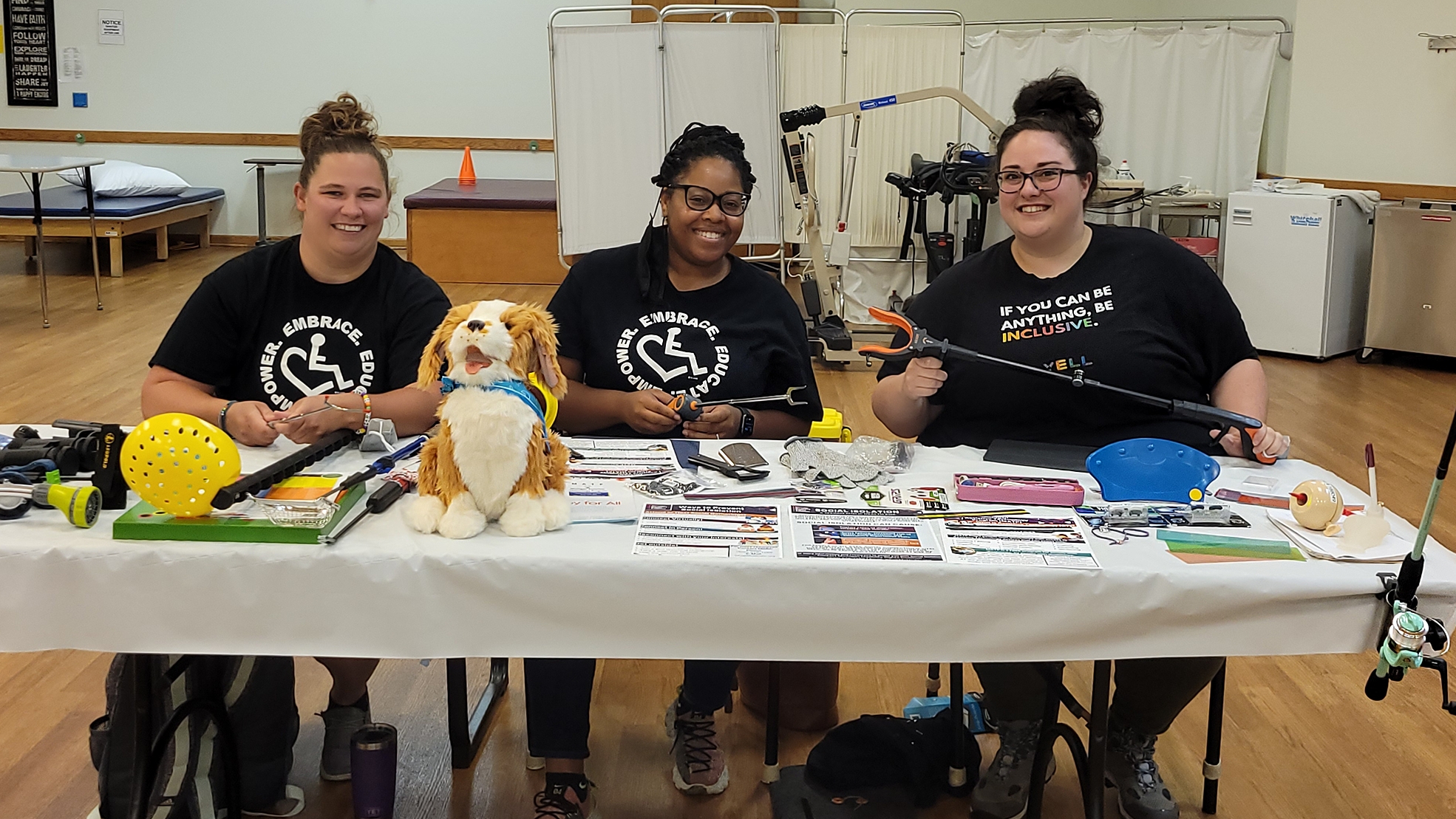 photo of a white woman with dark blonde hair up in messy bun next to her is a woman of color with black hair up in bun and next to her is a white woman with dark brown hair up in messy bun all three of them are sitting behind a table with an assortment of assistive technology on it