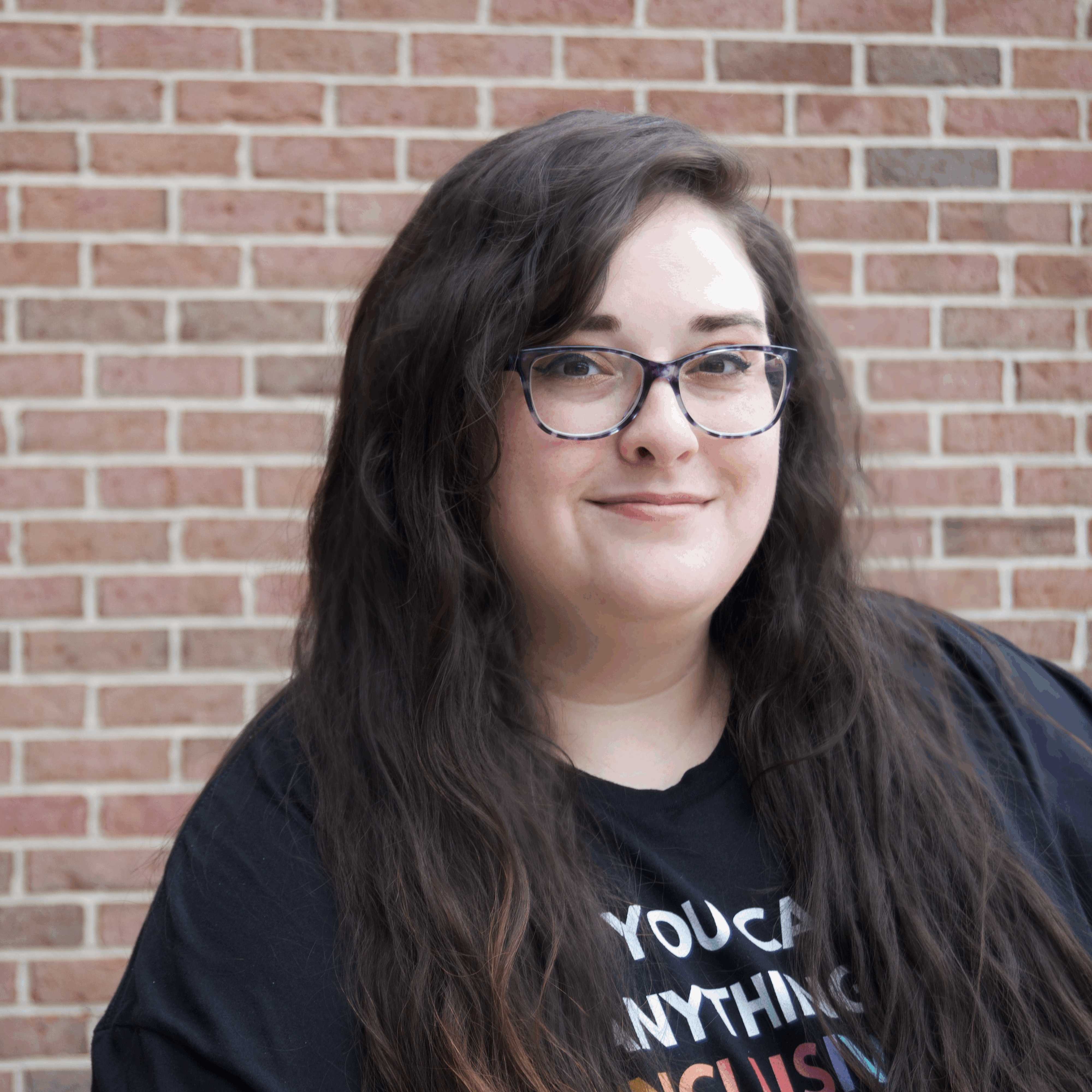 Abby White woman of size, smiling with long wavy dark brown hair. Dark rimmed glasses and black t-shirt in background of photo is brick background