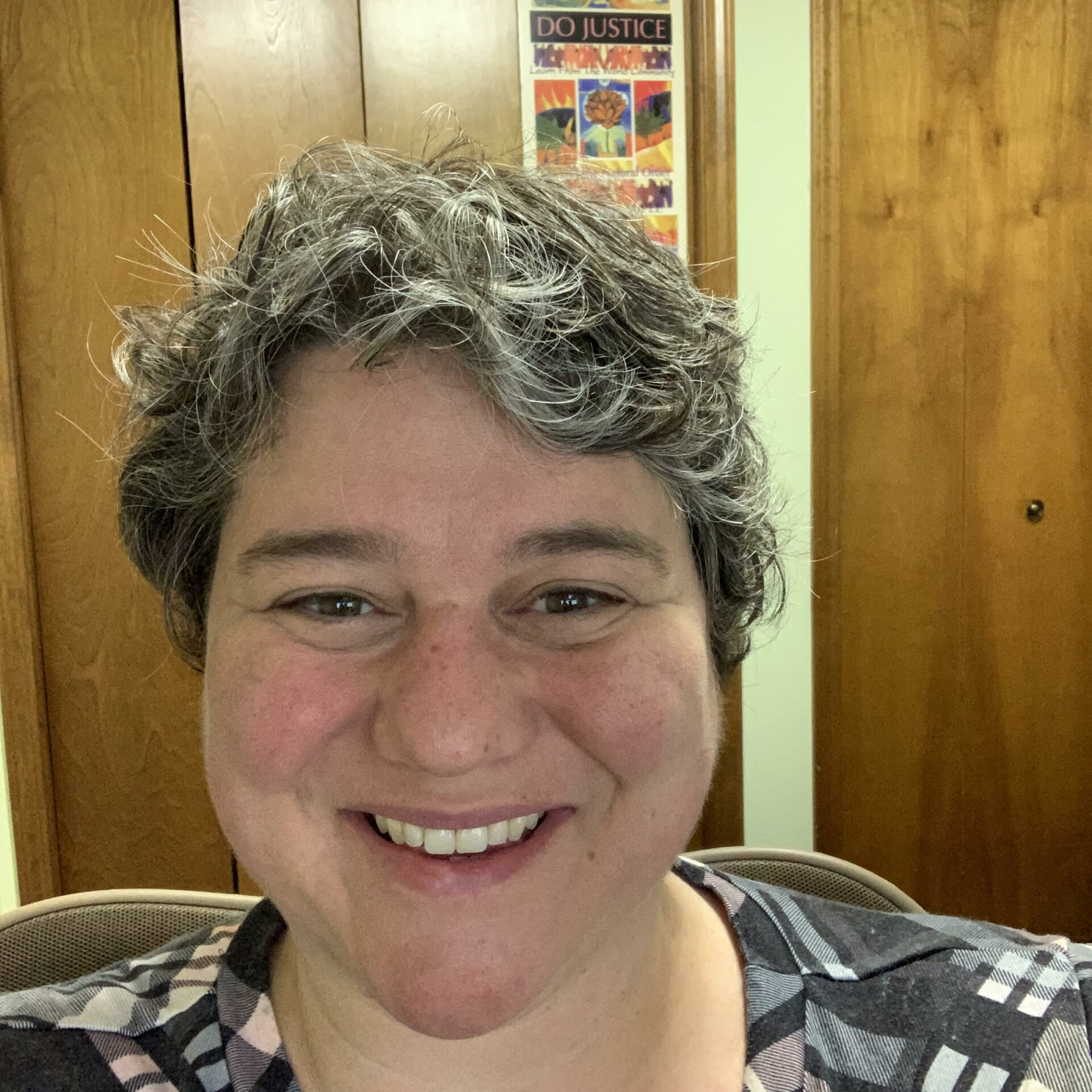 Aimee An image of a white woman with dark brown and gray curly short hair. The image is a selfie. She is sitting in her home office with a poster behind her that says "Do Justice'' she is wearing a black plaid vneck shirt.