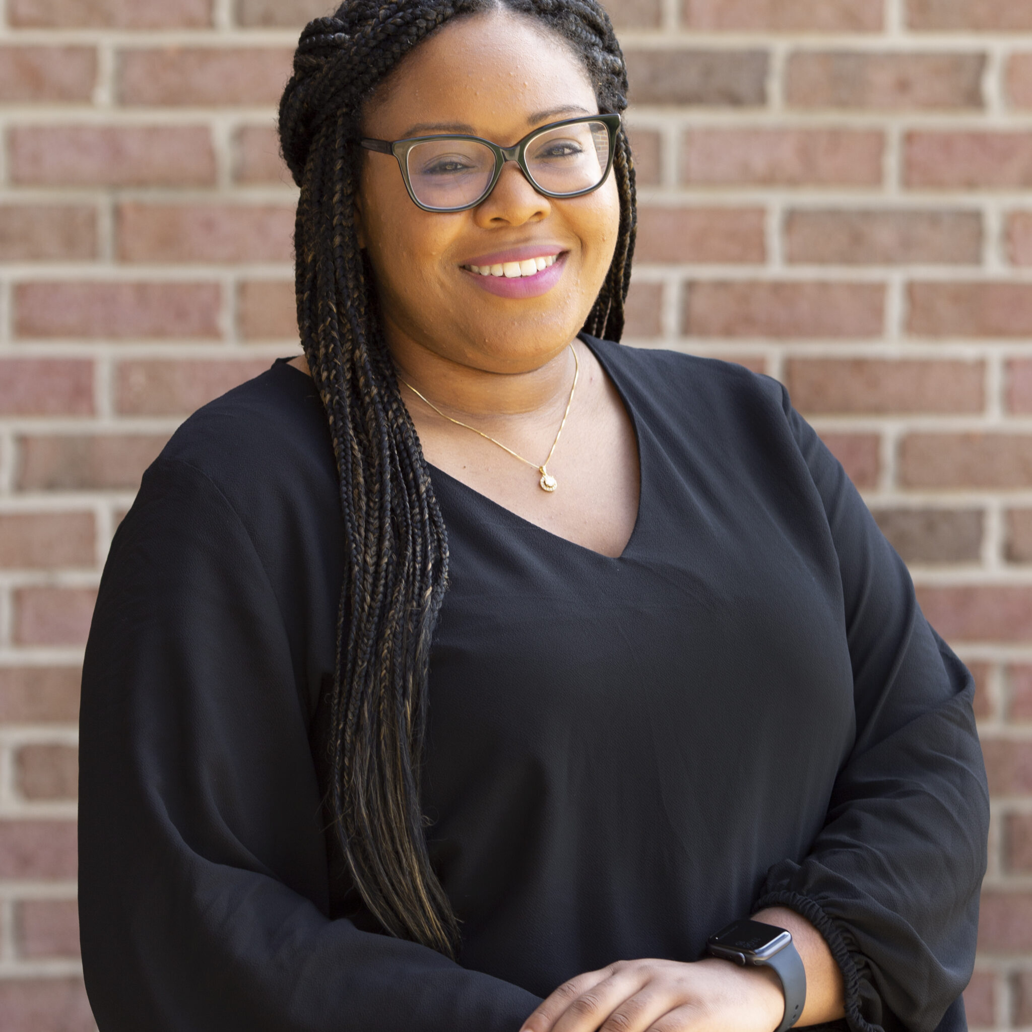 Ajaunè A woman with brown skin and long dark brown braids positioned with her hands crossed and smiling while standing in front of a brick wall. She is wearing black glasses and a black long sleeve blouse.