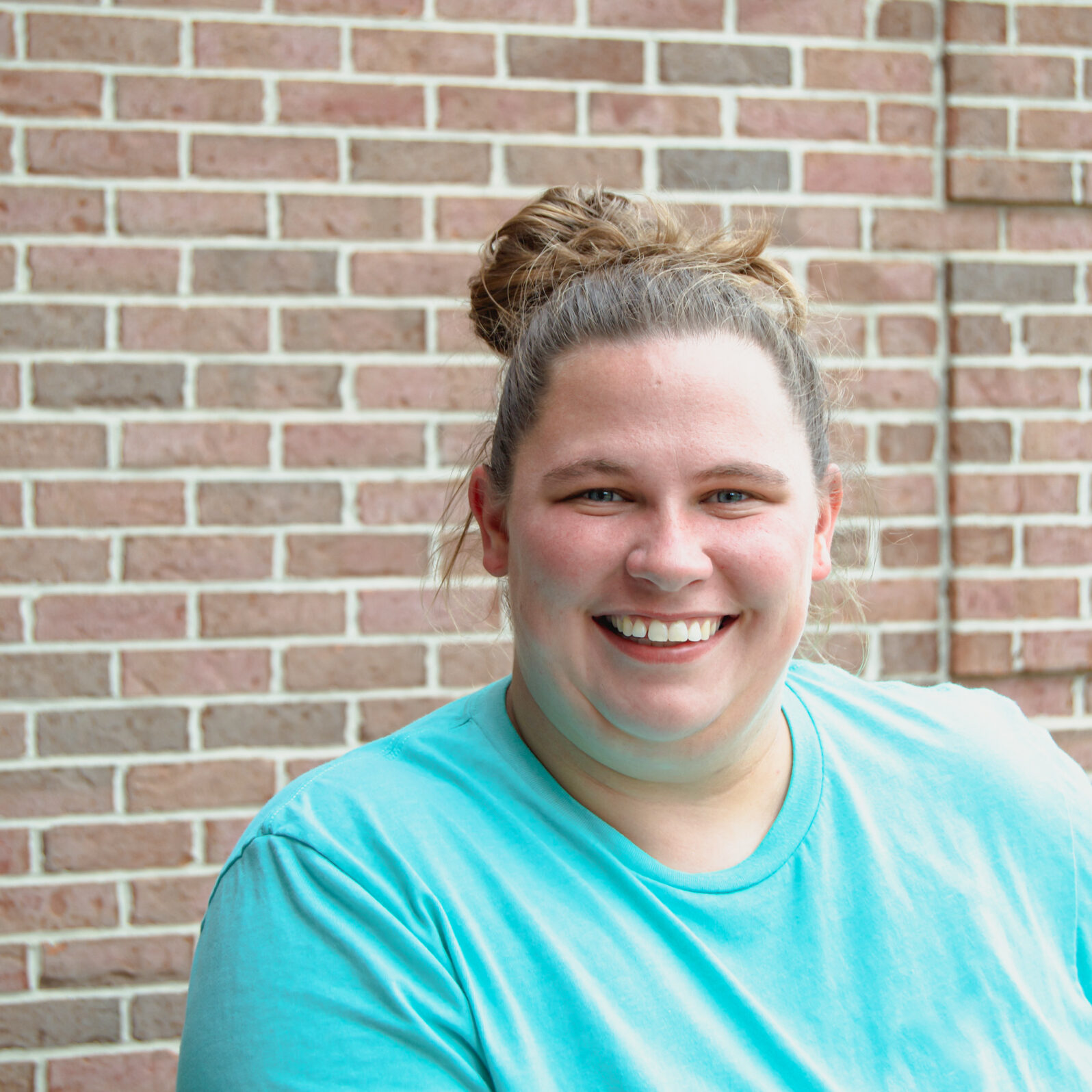 Jileesa White woman smiling with hair in a messy bun, with a light blue shirt on, standing in front of a brick wall