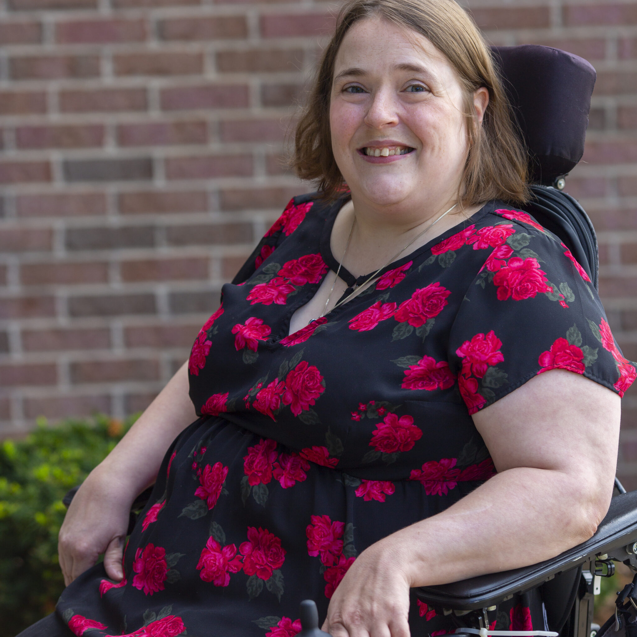 Laura A person with pale ivory skin and dark blonde hair smiling while sitting in a power wheelchair. They are wearing a black blouse with a pattern of red roses.