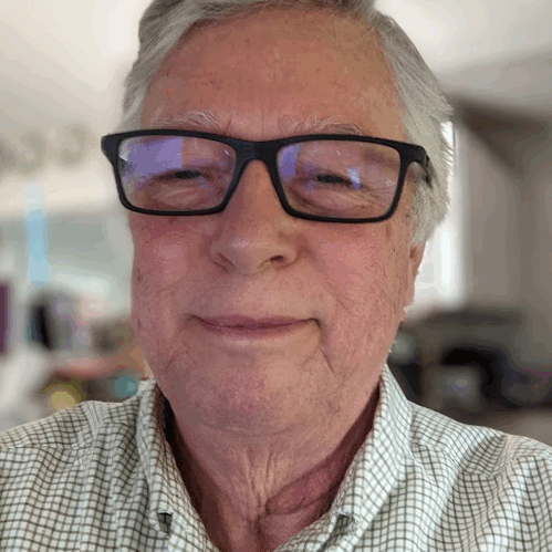 NormDelisle Norman DeLisle, elderly white male, with short gray hair, wearing a blue checkered shirt with suspenders; slight smile. He is sitting in his computer office at home, with part of his home kitchen in the background.