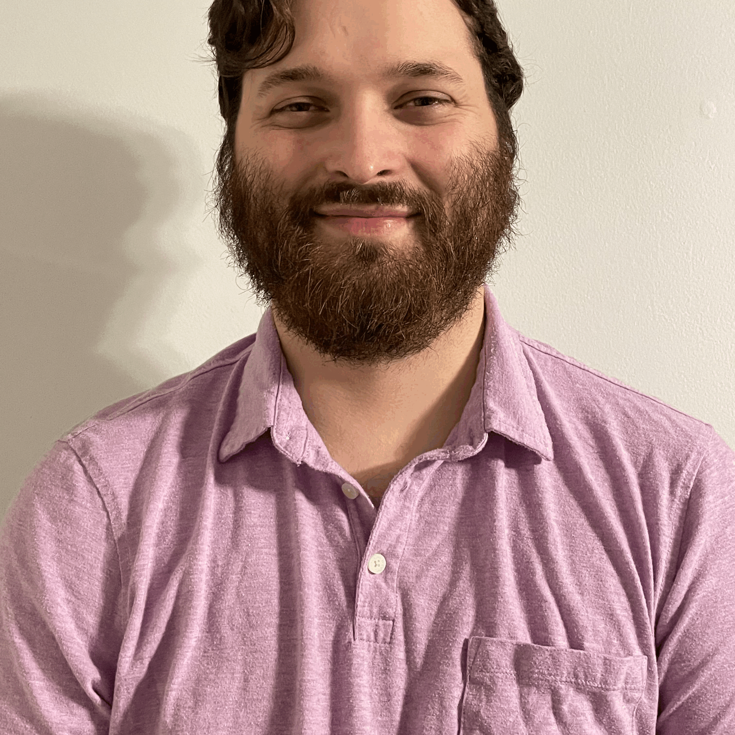 [Image description: Headshot of Brandon Taylor, a white man with short brown hair and a beard. Brandon is standing inside, with a blank white wall behind him. He is looking directly at the camera, smiling, and is wearing a light purple polo shirt. End image description.]